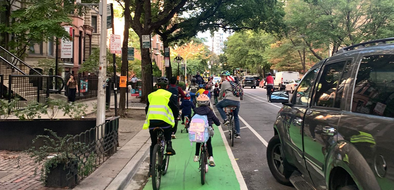 Families riding single file up the green protected bike lane, an adult in a hi-vis vest at the back